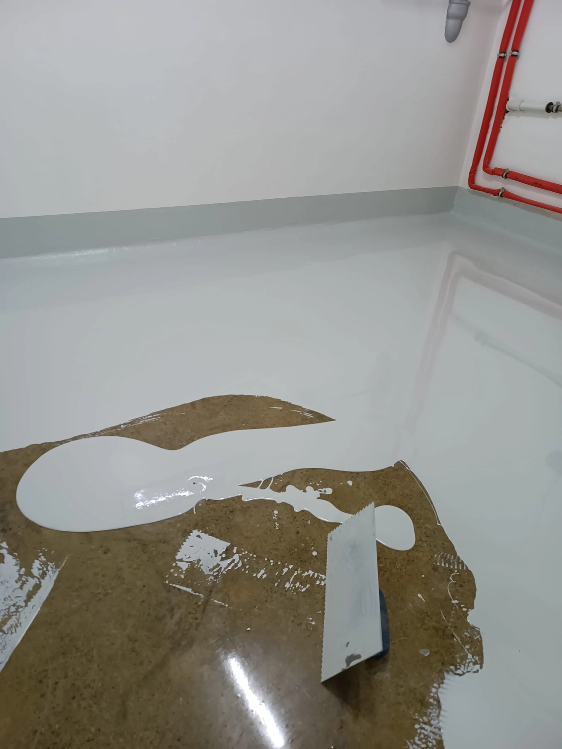 A&S Armbruster & Sieber GmbH - Experten im Bereich Bautenschutz und Bodenbeschichtungen - Auftragen einer weißen Epoxidbeschichtung auf einen Betonboden mit einer Zahnkelle in einer Ecke mit roten Rohren an der Wand.
